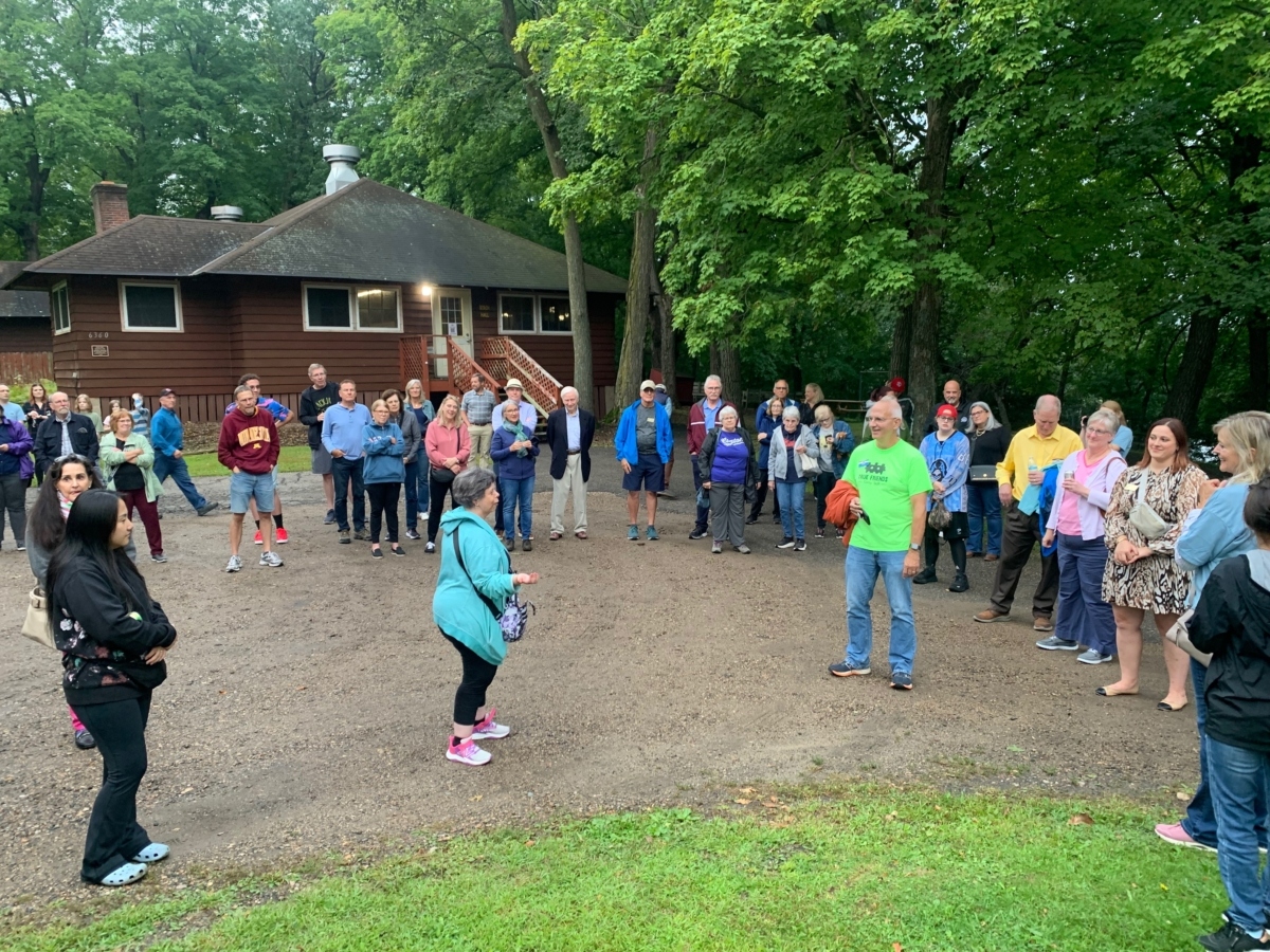 People arc around 4 others outside near woods and camp building.