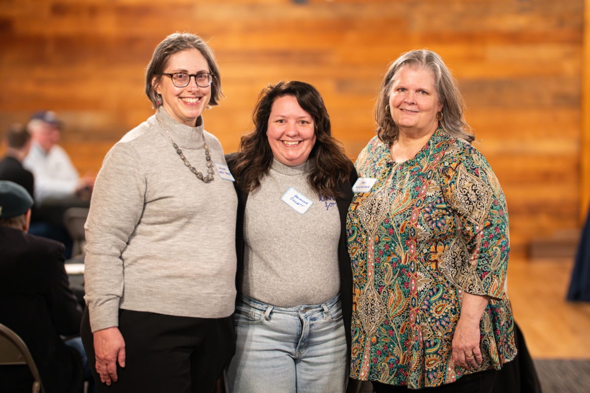 3 women pose in front of interior wood paneled wall