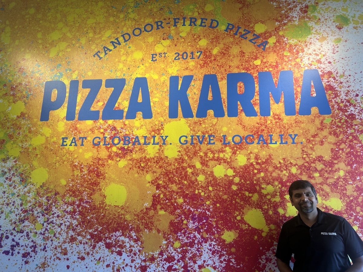 A man in a black shirt stands in front of a wall covered in splatters of yellow, orange and red paint. "Tandoor-Fired Pizza, Est. 2017, Pizza Karma, eat globally. Give Locally." is on top of of the paint splatter in blue paint.