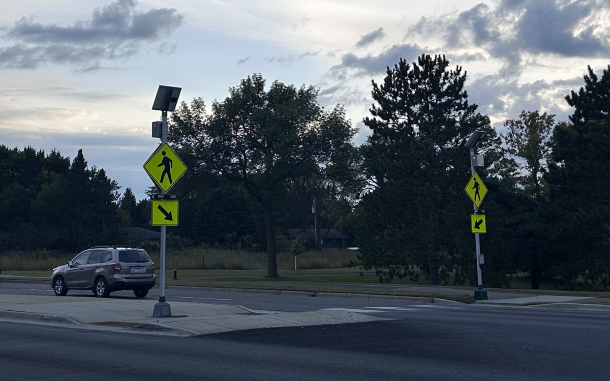 Two neon yellow road signs with pedestrians and arrows pointing to a crosswalk sit on a street.