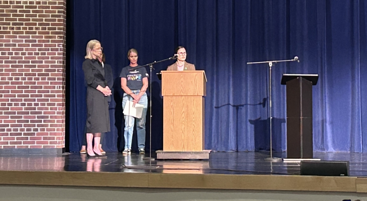 Three women stand on a stage in an auditorium with a full audience. One woman in a brown blazer stands behind a lectern.