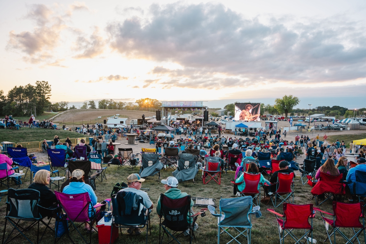 A large group of festival attendees sitting in lawn chairs watch a music performance in front of a sunset.