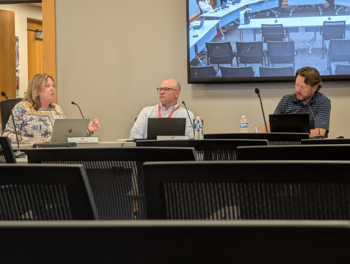 A woman speaks into a microphone at a school board meeting, while two other board members listen on. The board members are sitting at a table with laptops in front of them.