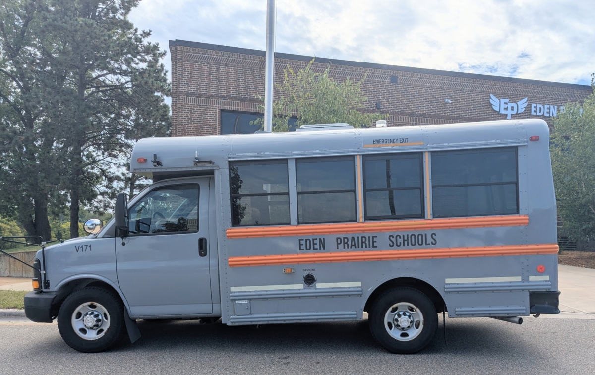 A blue and orange school bus in front of the Eden Prairie Schools administrative building. The bus says "Eden Prairie Schools" on the side.