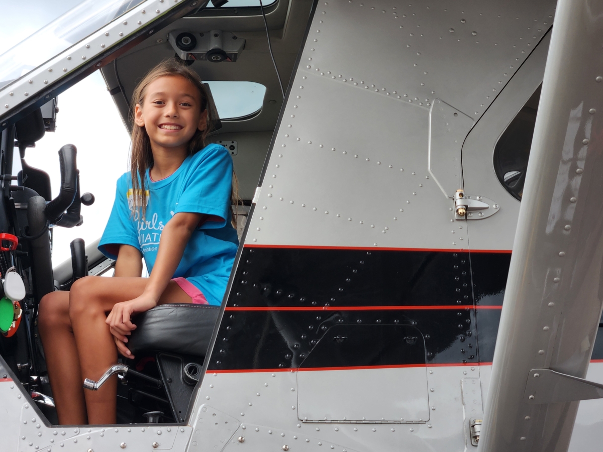 young girl in blue T-shirt sits and smiles in pilot's seat of grounded airplane