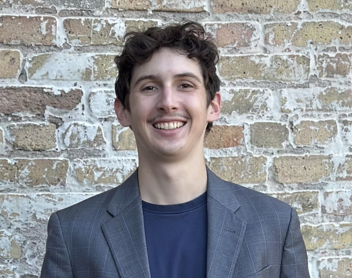A man in a checked suit jacket and a navy t-shirt smiles in front of a brick wall.