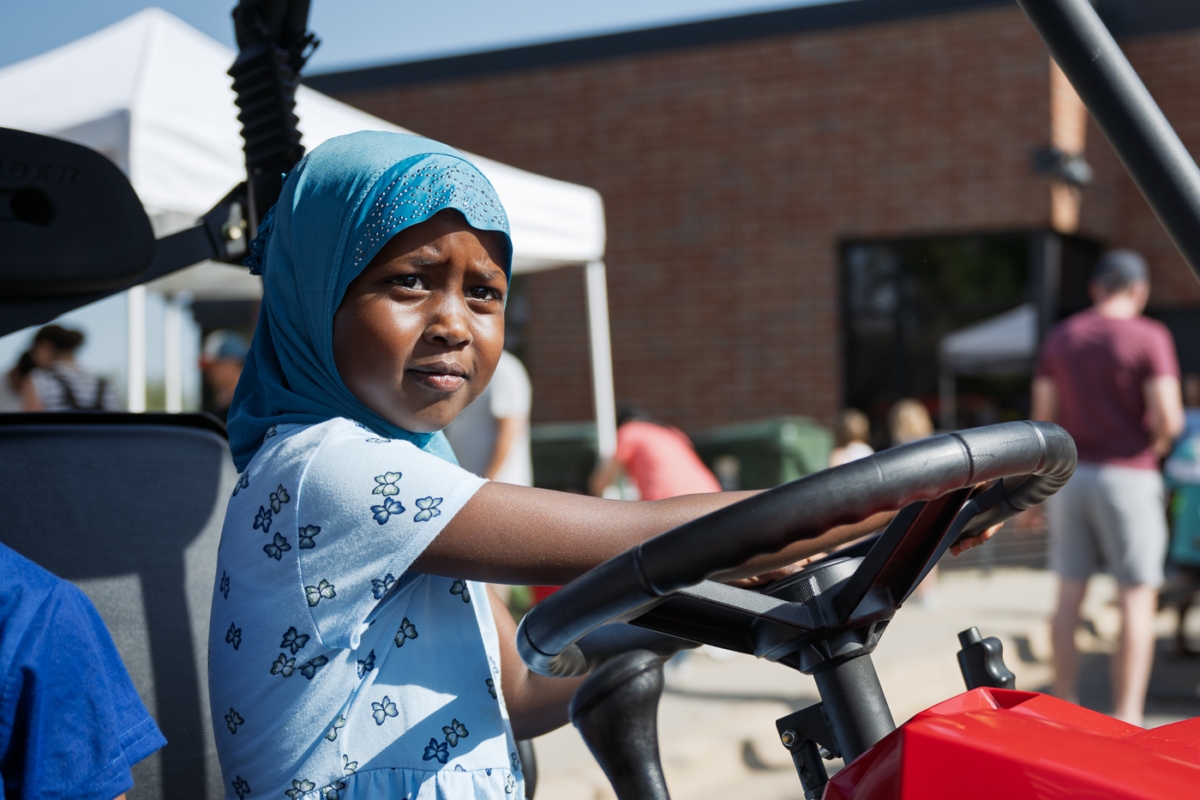 Young girl wearing blue hijab sits at steering wheel of utility vehicle at outdoor community event