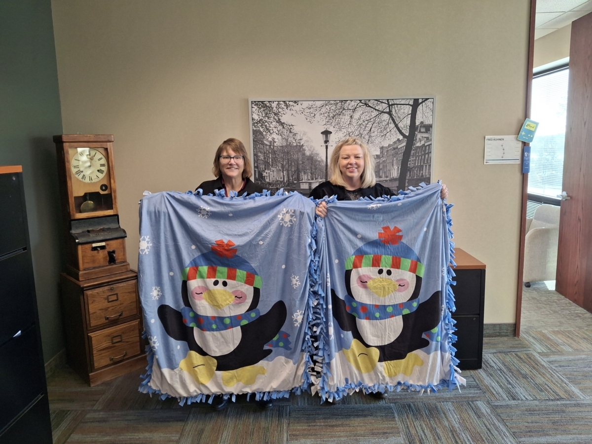 2 women stand and pose with blue tied blankets with large images of penguins on them