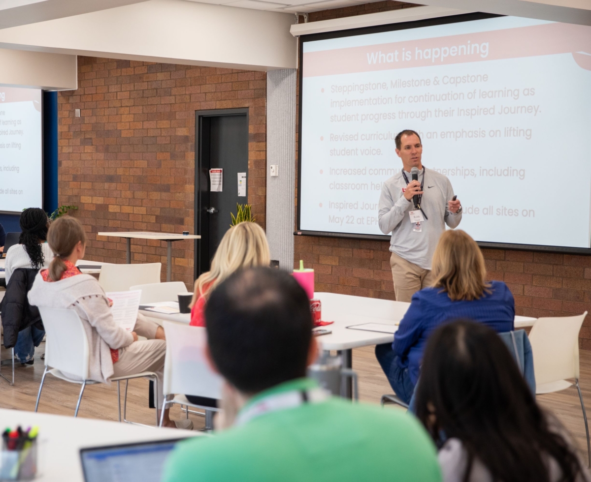 A person with a microphone speaks to a crowd of people sitting at white tables. A presentation is projected onto the screen behind the speaker.