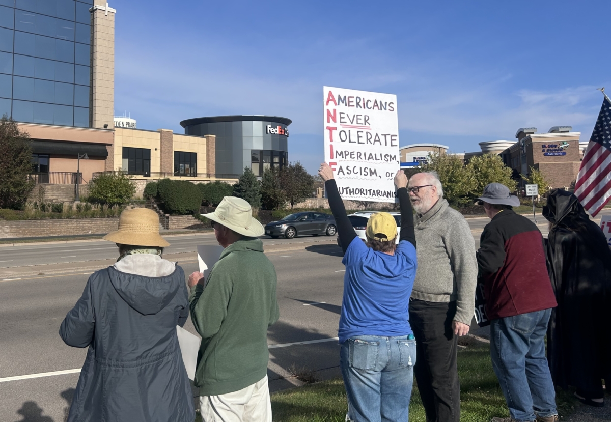 A woman stands in a group of people on the side of the road. She holds a sign that says "Americans Never Tolerate Imperialism, Fascism, or Authoritarianism".