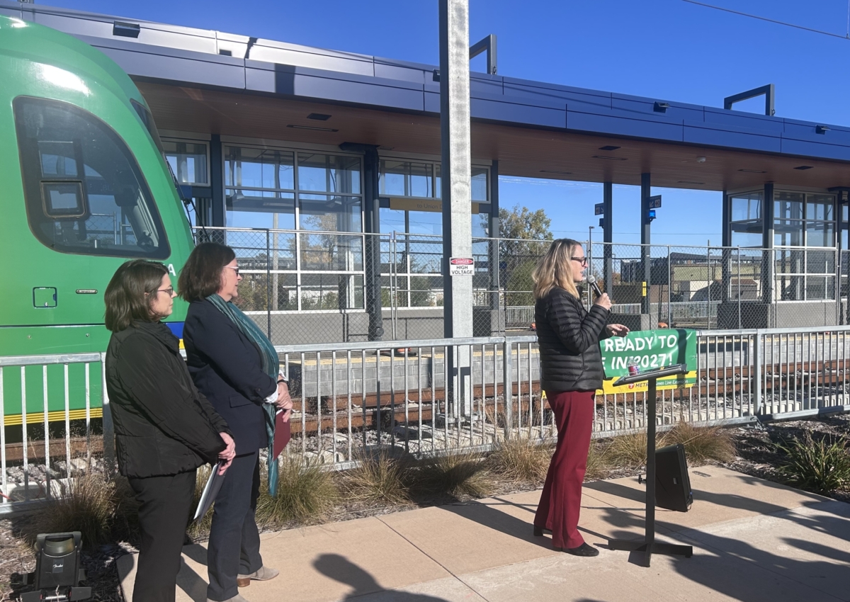 Three women stand in front of a green light rail car and light rail station. One woman holds a microphone