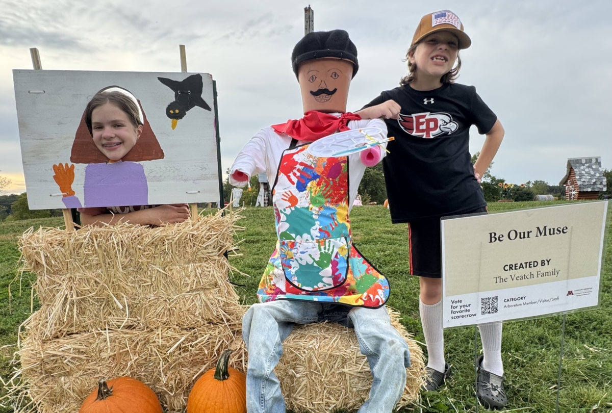 Two kids stand beside a scarecrow. The scarecrow is wearing an apron and holding a painter's palette and brush. On its left, a girl is peaking from behind a canvas painting and on its right a boy is leaning against it.
