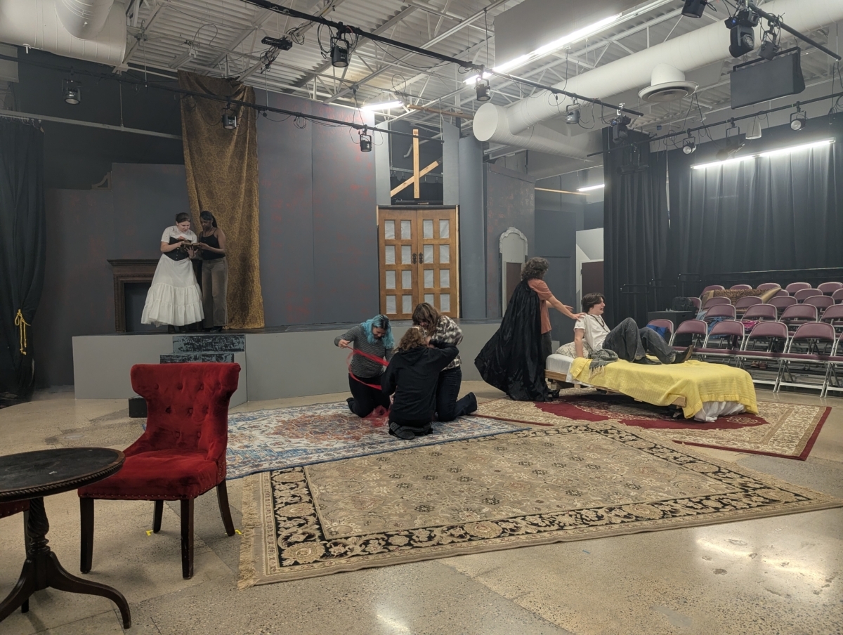 Students in various period costume pieces practice "Dracula." They are in a theater with a small stage, a chair and table, a bed, and some rugs. Two students are reading a book on the stage, three students are miming with red ribbon in the middle of the floor, and one student is sitting on the bed with another student standing above them.