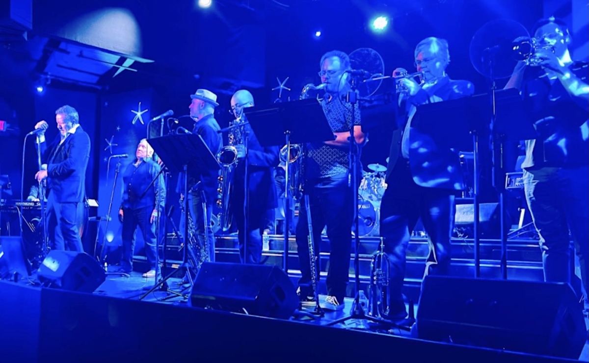 Musicians perform on stage during “Spinning Wheel Trilogy” at Chanhassen Dinner Theatres, featuring a full horn section under blue stage lights.