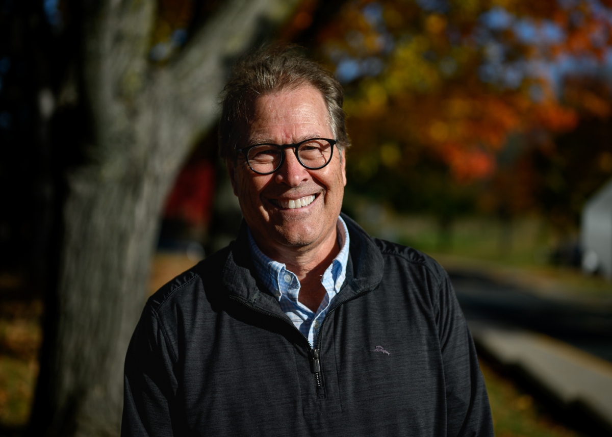 A man wearing glasses and a black coat stands in front of a tree bearing red and orange leaves.