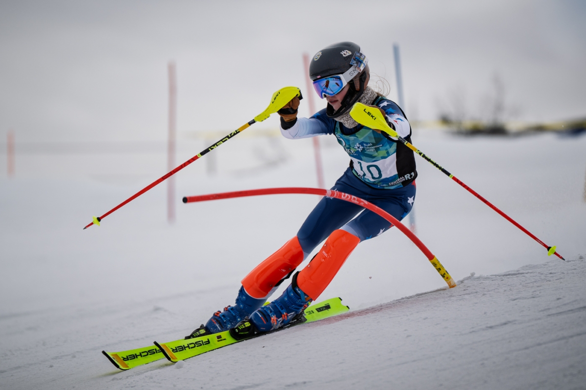 A female skier in a blue racing suit and orange leg guards makes a turn through a slalom course on a snowy slope with gates visible in the background.