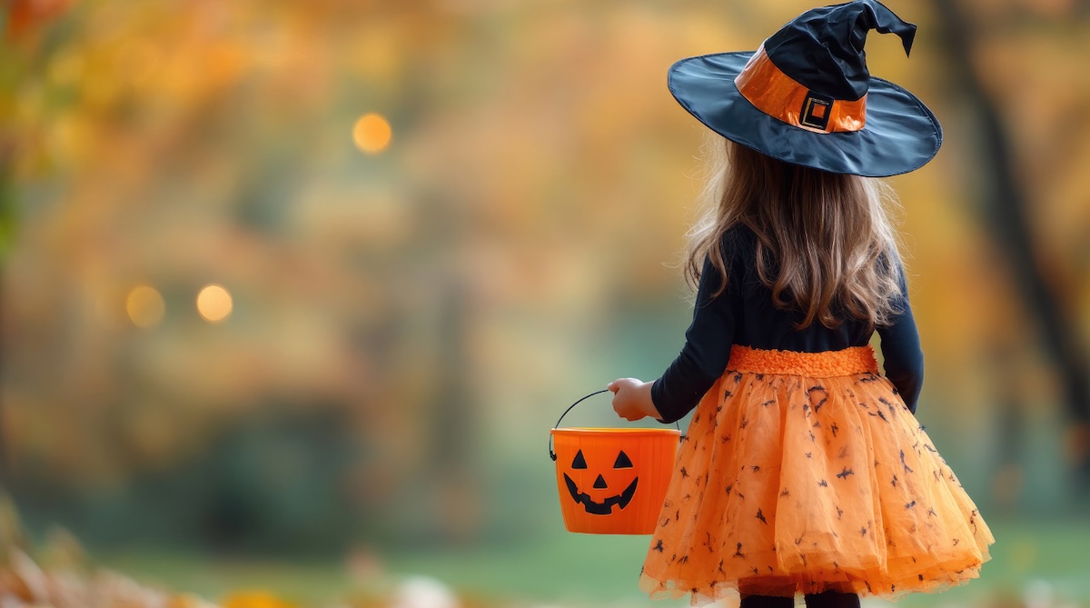 A young girl dressed as a witch holds a pumpkin-shaped bucket while trick-or-treating on a fall evening.