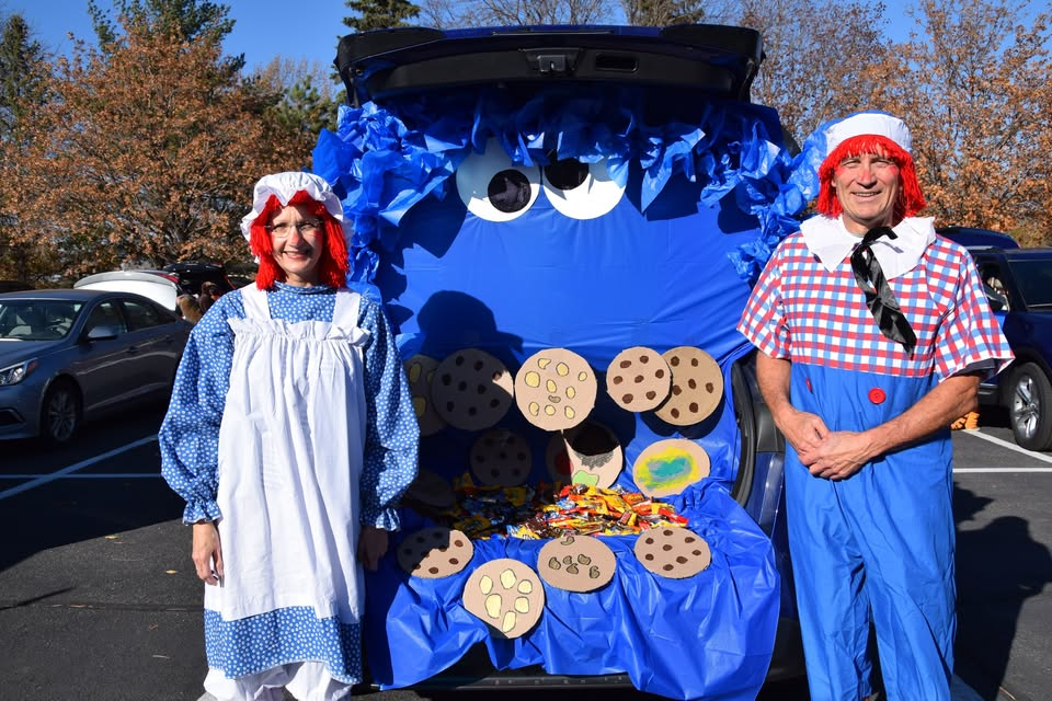 Two adults dressed as Raggedy Ann and Raggedy Andy standing next to a car decorated as Cookie Monster