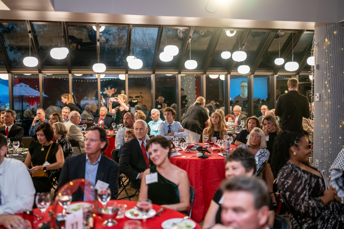Eagle Excellence Showcase attendees watching the program. The tables have red tablecloth and have plates of food and glasses on them.