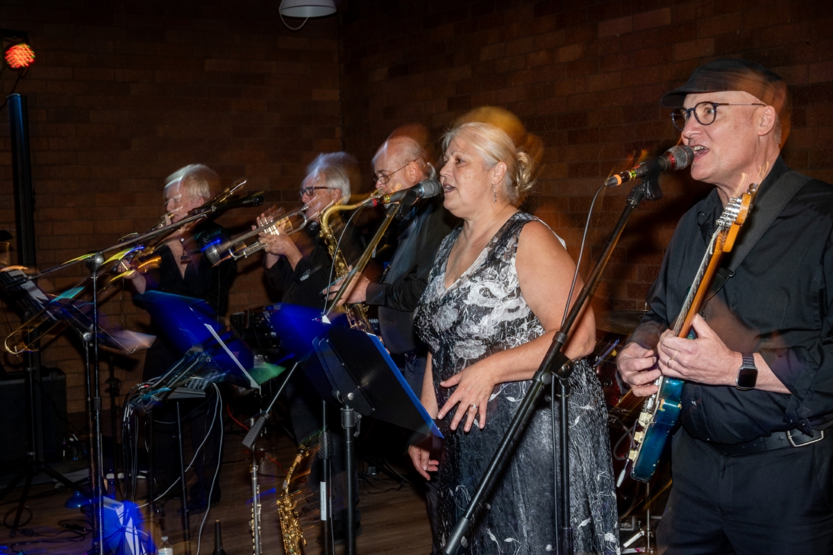 Five musicians performing. Two of the performers are singing into microphones, with one of them holding a guitar. One of the other performers is playing a saxophone, and the other two are playing trumpets.