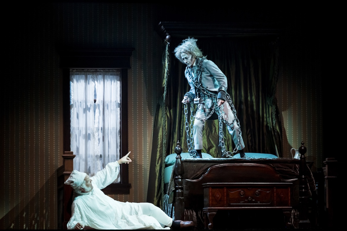A ghostly figure draped in heavy chains stands on a canopy bed, confronting a frightened man in a nightcap and nightshirt who points upward from where he lies. The scene is dimly lit and staged inside a Victorian bedroom with striped wallpaper and a curtained window.
