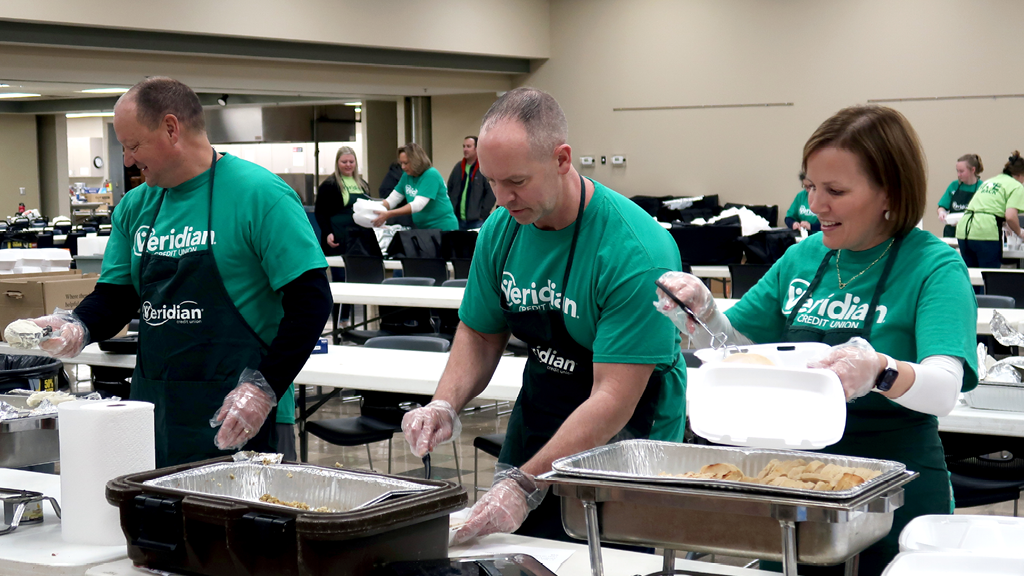 3 people in green T-shirts and black aprons dishing up food in a cafeteria setting