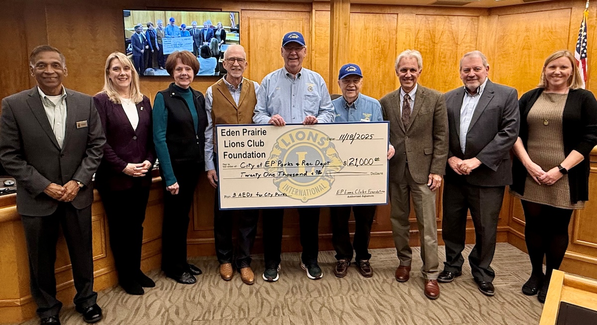Eden Prairie City Council members and Eden Prairie Lions Club Foundation representatives stand in the council chambers holding a large ceremonial check for $21,000 donated for new public-access AEDs in city parks.