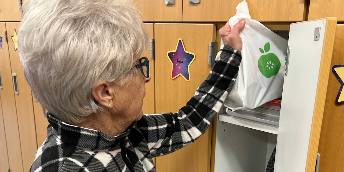 An older woman puts a white Every Meal food bag into a student locker labeled “Ana” at an elementary school.