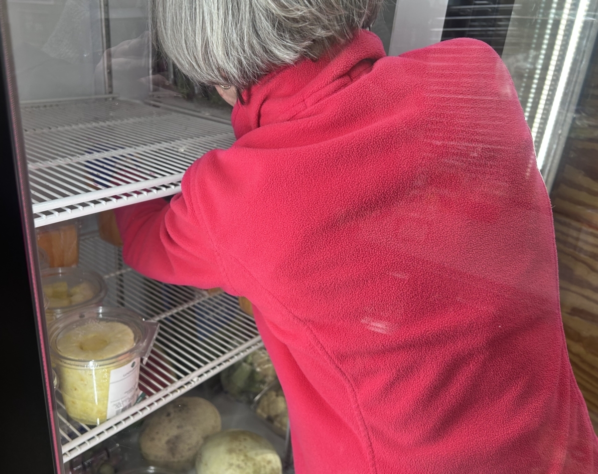 A woman in a red sweat shirt stocks a produce refrigerator with fruit.