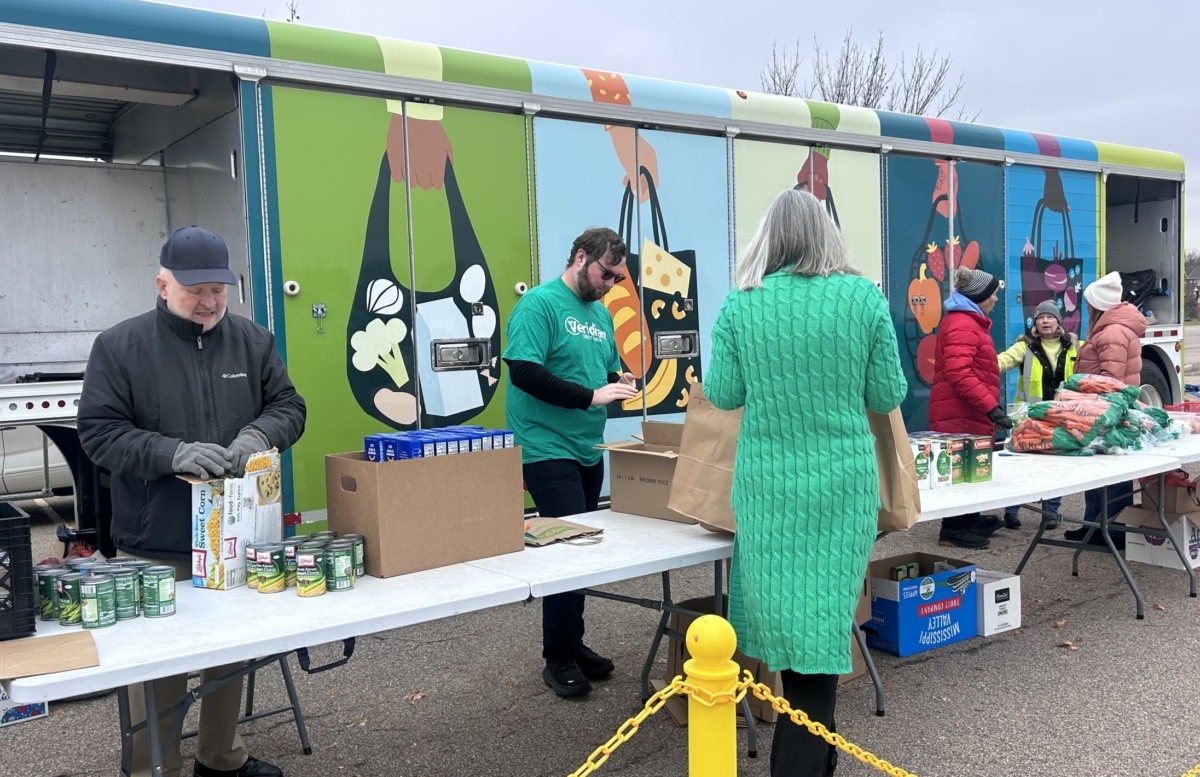 A truck decorated with people holding grocery backs full of food sits behind a group of volunteer distributing canned goods and boxes of pasta.