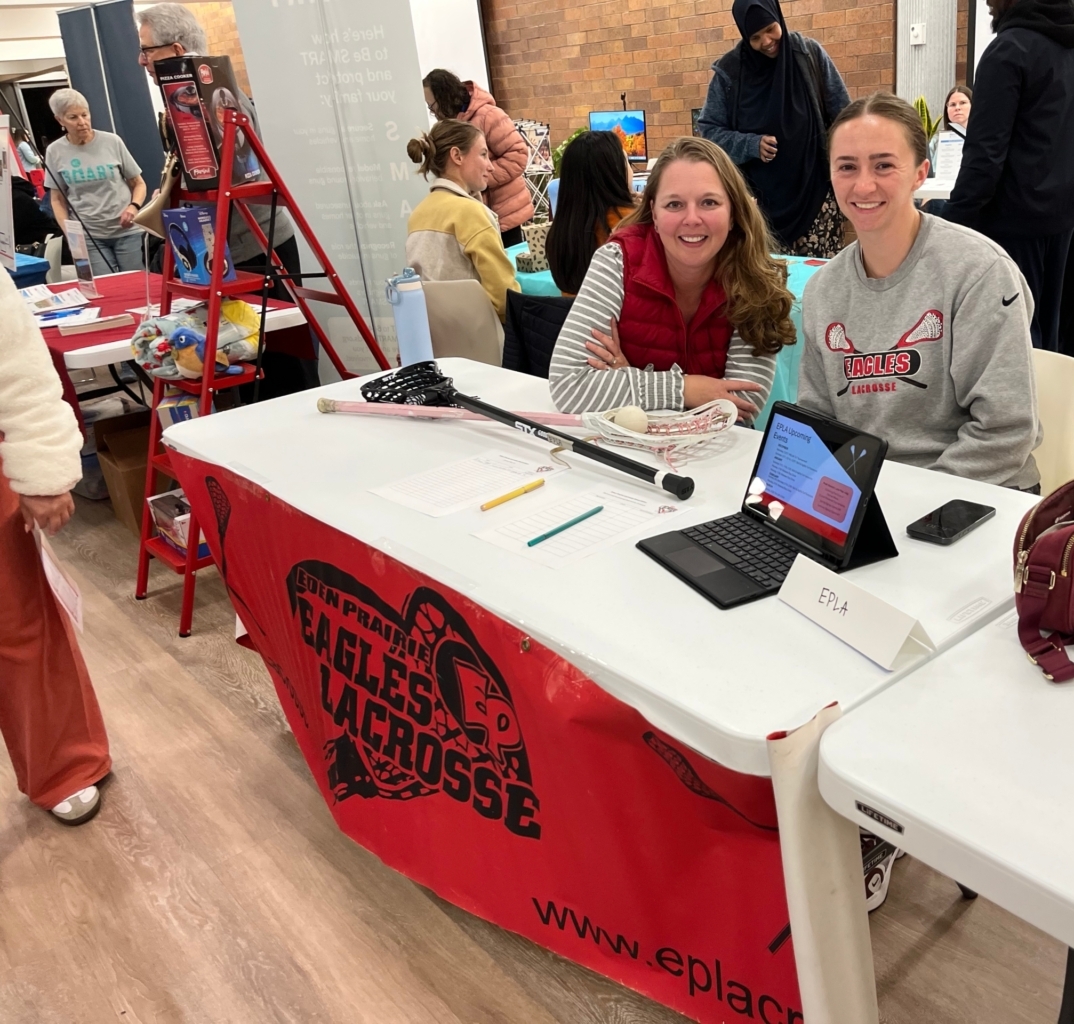 2 women smile while sitting behind a table with a lacrosse stick on top of it