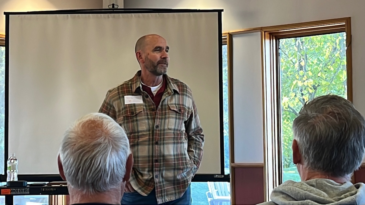 Doug Boonstra speaks to an audience during an indoor event, standing near a projection screen and windows.