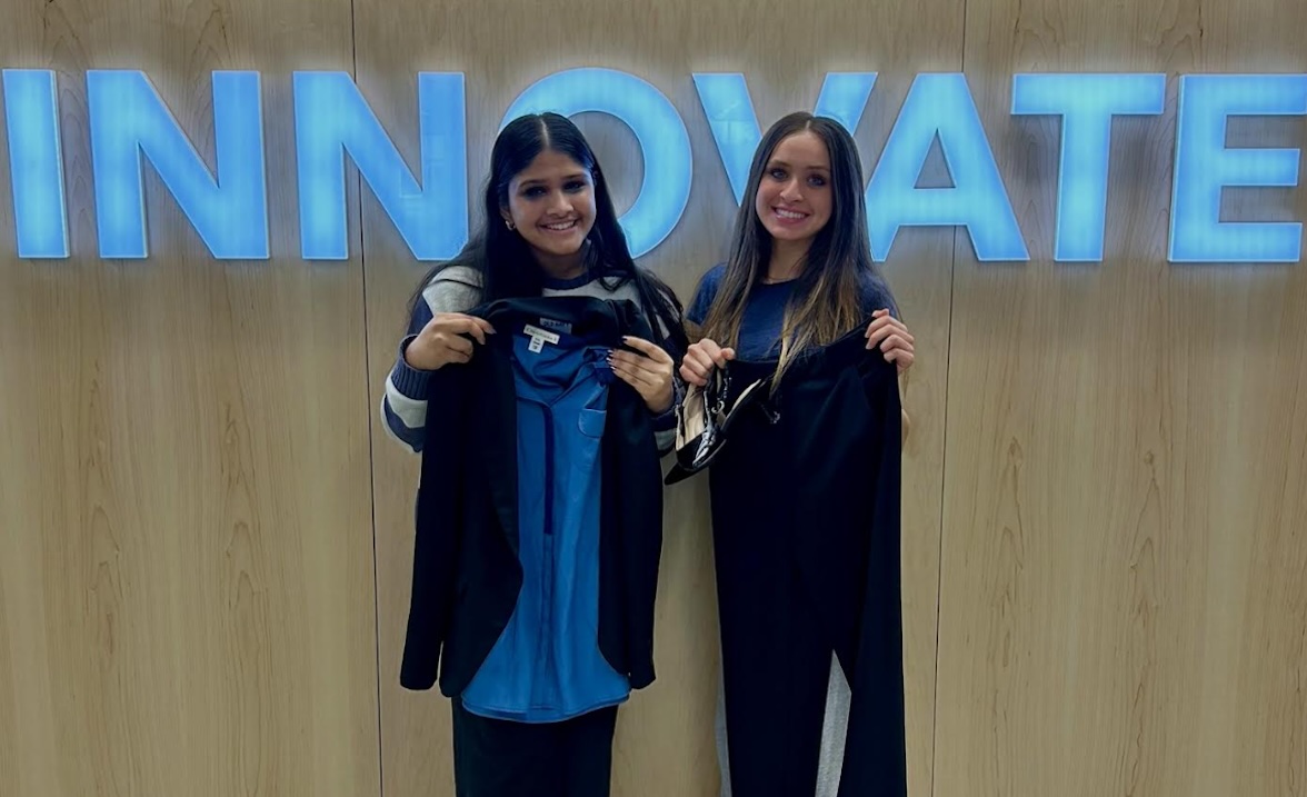 Aishah Alam and Avni Maheshwari smile while holding business professional clothes for the LeadHER clothing drive. The word "Innovate" is in glowing, blue letters behind them.