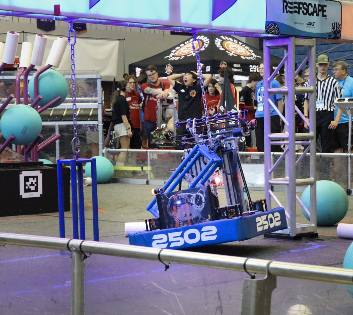 blue robot approaching green balls as people observe in background during robotics competition