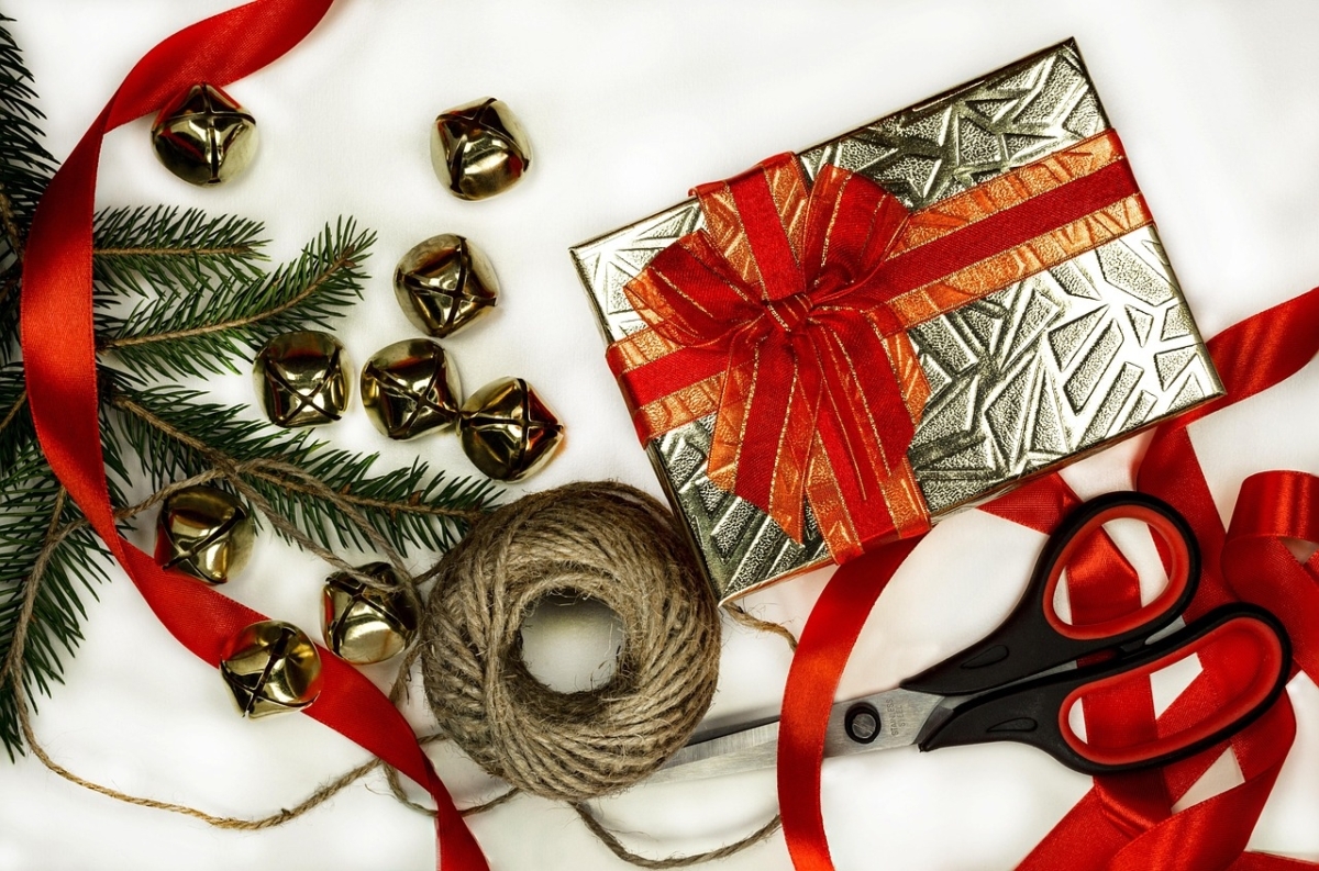 A photo of a gift wrapped box with a red ribbon and bow, surrounded by more ribbon, string, scissors, small jingle bells, and some pine tree boughs.