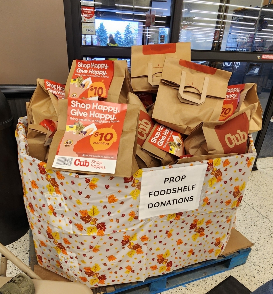 A large box decorated with a fall theme, containing many purchased bags of groceries donated by customers.