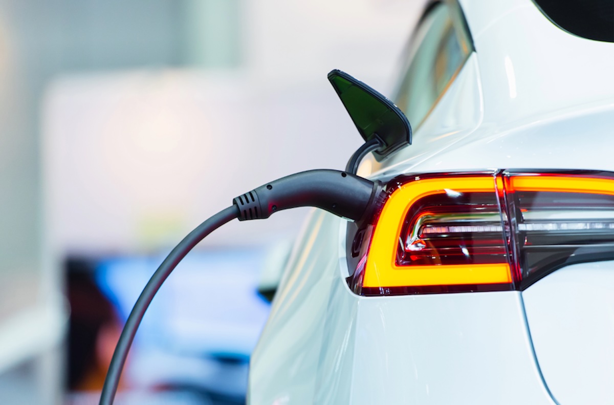 An electric vehicle charges at a public charging station, with the charging cable connected near the rear taillight.