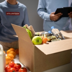 A photo of a woman wearing a blue shirt with the word "volunteer" on it, standing behind a table with a brown cardboard box filled with food.