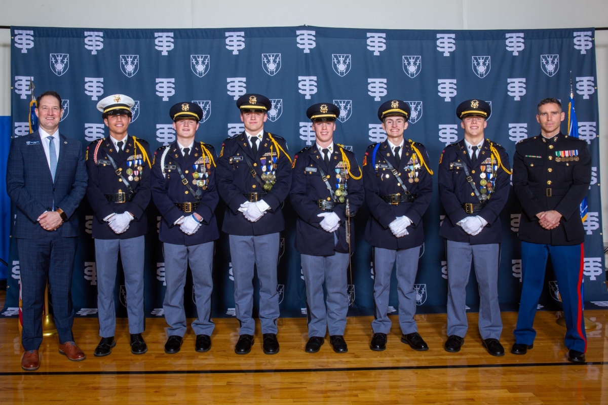6 young men in military uniforms stand between 2 older men, 1 in suit and 1 in uniform