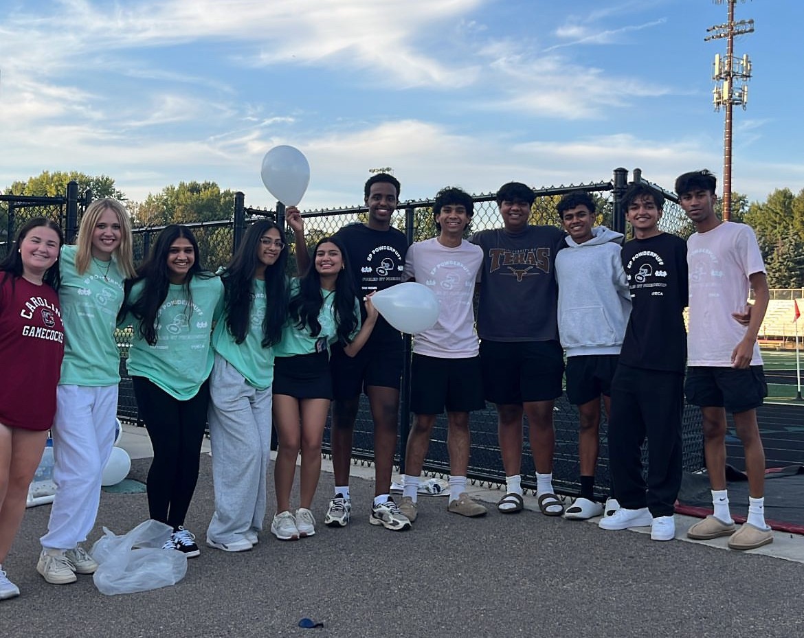 Eleven Eden Prairie High School students smile for a photo in front of the school's football field. A few of them are holding white balloons, and a fence is behind them.