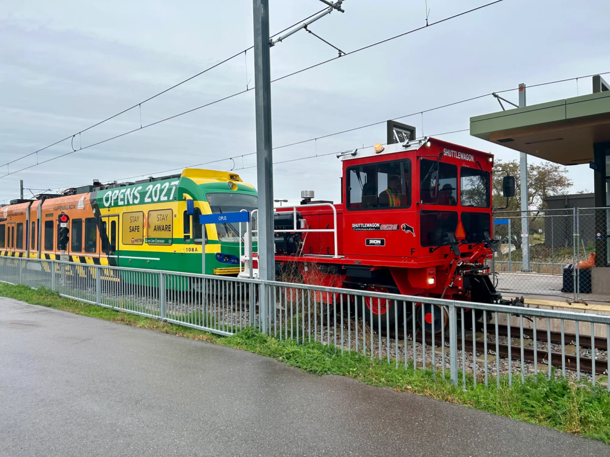 A Green Line Extension light-rail train is towed by a red maintenance vehicle along the tracks.