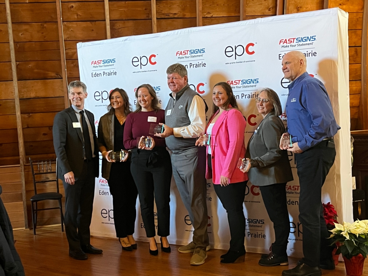group of people holding clear glass awards stands in front of sign with Eden Prairie Chamber of Commerce logo