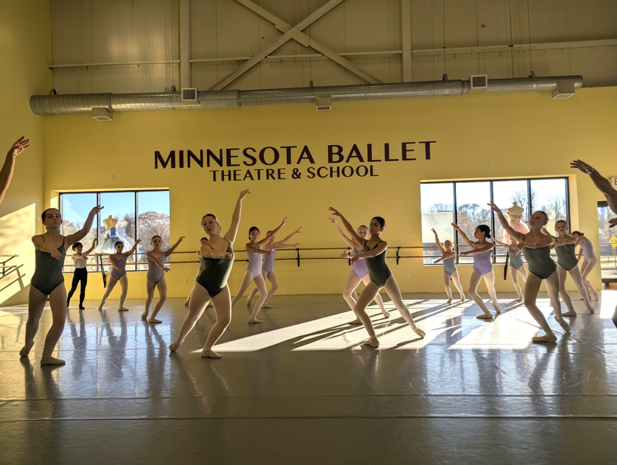 Dancers in leotards strike a ballet pose. The walls are yellow and the words "Minnesota Ballet Theatre & School" are written in large letters. There are two windows with ballerina costumes displayed, along with ballet barres underneath.