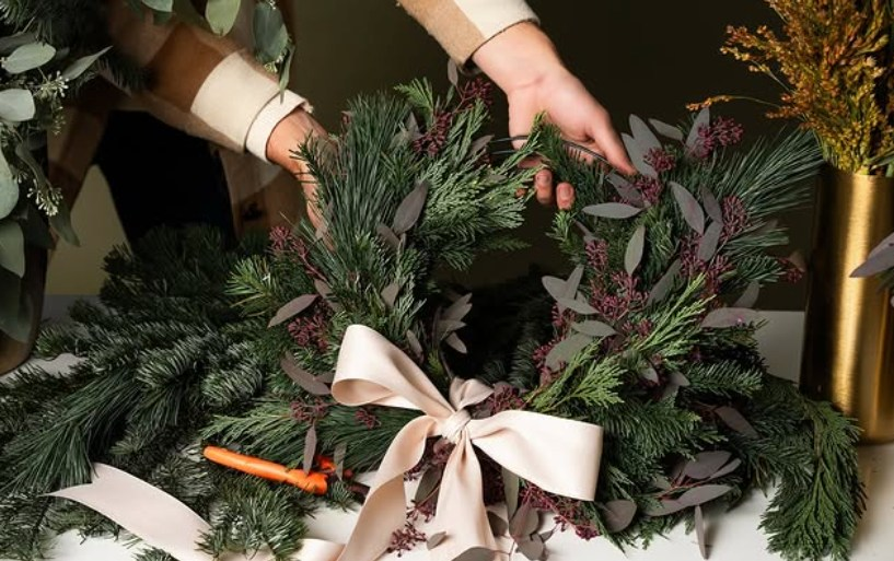 A wreath of evergreens with a white bow at the bottom being held up by the hands of a woman wearing a sweater.